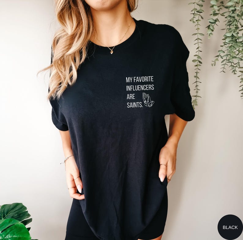 May include: Black t-shirt featuring the white text "MY FAVORITE INFLUENCERS ARE SAINTS." and a graphic of praying hands. The t-shirt has a crew neck design.