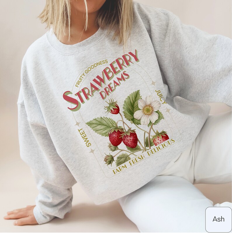 May include: A gray sweatshirt with a graphic print of strawberries, flowers, and the text "Strawberry Dreams" in a vintage style. The text also includes the words "Fruity Goodness", "Sweet", "Juicy", "Farm Fresh", and "Delicious".