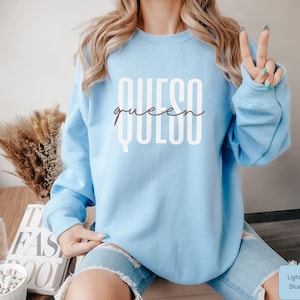 May include: Light blue sweatshirt with the text "QUESO queen" in white lettering.