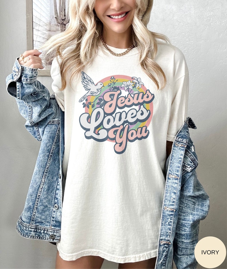 A model is wearing an ivory colored shirt with a graphic that says Jesus Loves You in a retro font script and there is a circulat rainbow behind with a white dove and some lilies. The colors are retro-muted colorful colors.