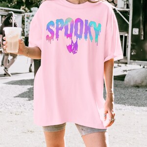 May include: A light pink t-shirt with the word "SPOOKY" in a dripping, rainbow-colored font. A bat graphic is below the word. The word "Blossom" is printed at the bottom of the image.