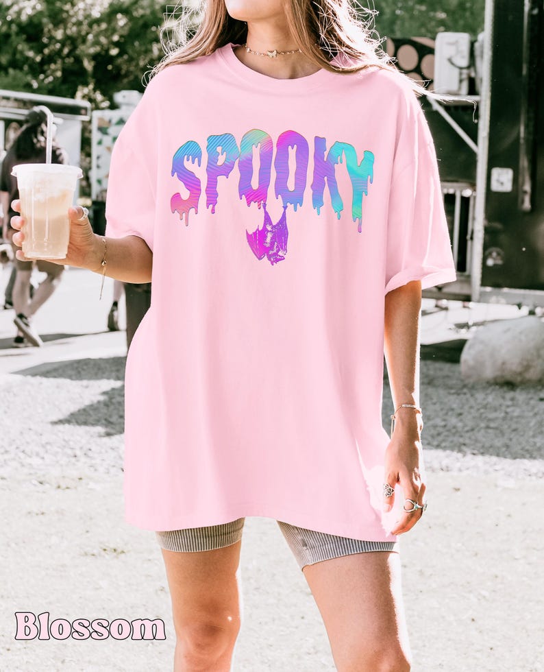 May include: A light pink t-shirt with the word "SPOOKY" in a dripping, rainbow-colored font. A bat graphic is below the word. The word "Blossom" is printed at the bottom of the image.