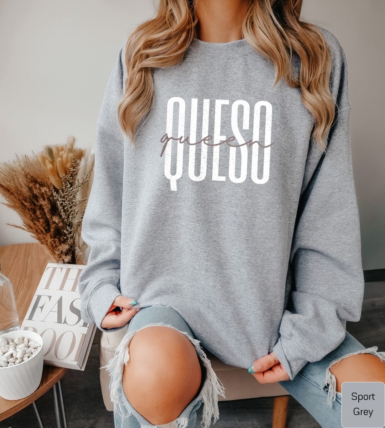 May include: A gray sweatshirt with the text "QUESO queen" printed in white on the front. The sweatshirt is being worn by a person with long blonde hair.