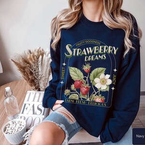 A model wears a navy sweatshirt with a vintage-inspired graphic of strawberries on the stem. It says Strawberry dreams in an art-deco font with other highlighted words around that say fruity goodness, sweet, juicy, farm fresh, and delicious.