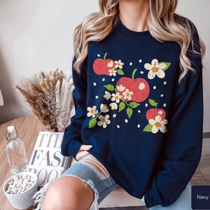 May include: A navy blue sweatshirt with a graphic of red apples and white flowers. The sweatshirt has a crew neck and long sleeves.