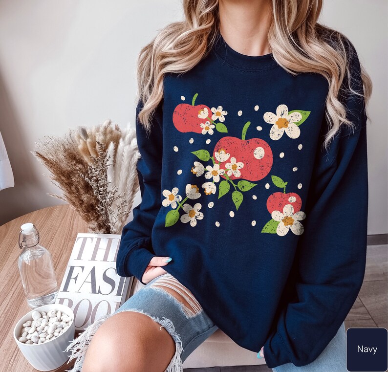 May include: A navy blue sweatshirt with a graphic of red apples and white flowers. The sweatshirt has a crew neck and long sleeves.