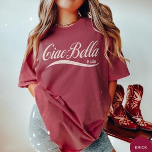 A model wears a brick shirt that says ciao-bella is a classic script style. It has a wavy ribbon underneath and says Italia.