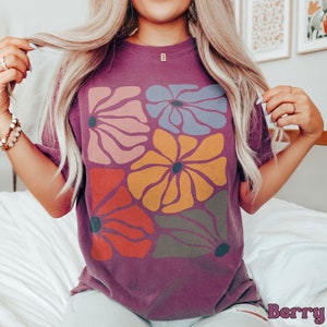 A model wears a berry shirt with abstract, wavy, boho flowers in colors of muted mauve, light grey-blue, gold, red, and dark taupe all with deep blue pistils. The colorful design is a slightly distressed texture.