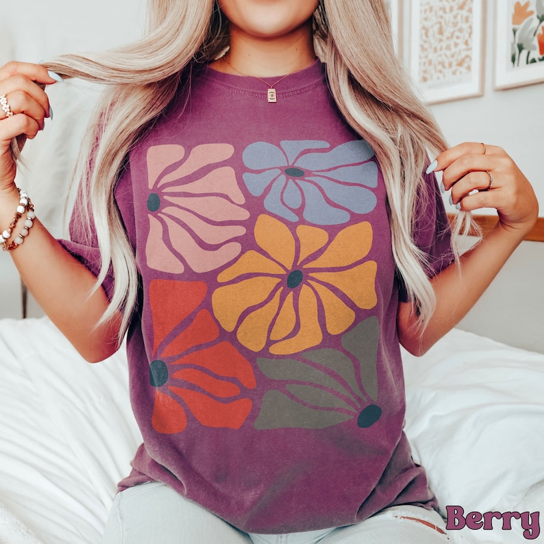 A model wears a berry shirt with abstract, wavy, boho flowers in colors of muted mauve, light grey-blue, gold, red, and dark taupe all with deep blue pistils. The colorful design is a slightly distressed texture.