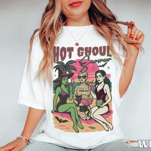 May include: White t-shirt with a colourful graphic that reads "HOT GHOUL SUMMER." The graphic features three cartoon figures: a witch, a mummy, and a vampire, all enjoying a beach scene. The witch is holding a mirror, the mummy is reading a book, and the vampire is holding a drink.