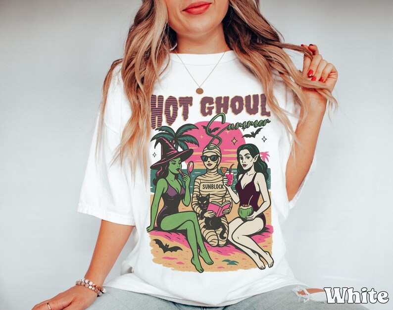 May include: White t-shirt with a colourful graphic that reads "HOT GHOUL SUMMER." The graphic features three cartoon figures: a witch, a mummy, and a vampire, all enjoying a beach scene. The witch is holding a mirror, the mummy is reading a book, and the vampire is holding a drink.