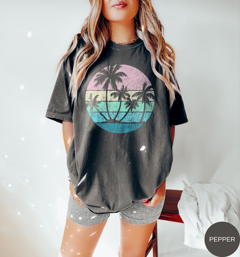 A retro sunset with faded and distressed colors of pink, yellow, green, and two shades of blue have some see through palm trees in front for a vintage look, on a pepper-colored shirt on a model.