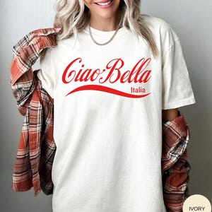 A model wears an ivory shirt that says ciao-bella is a classic script style. It has a wavy ribbon underneath and says Italia.