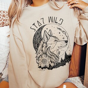 May include: A beige sweatshirt with a black wolf howling at the moon graphic. The graphic also includes a floral design and the text "Stay Wild".