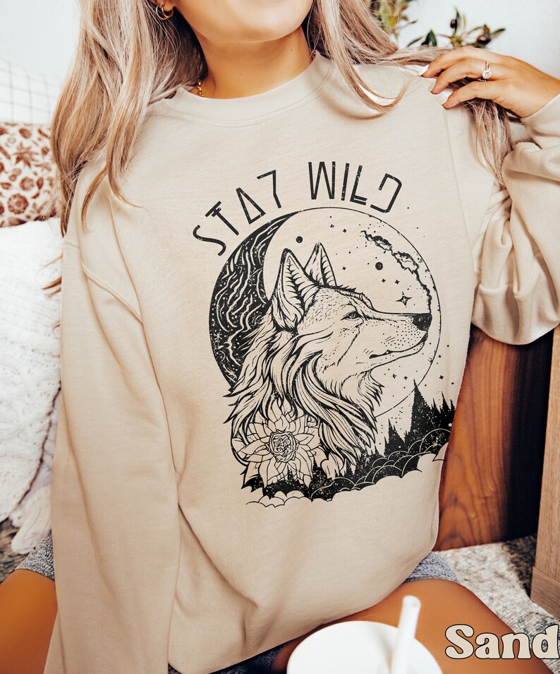 May include: A beige sweatshirt with a black wolf howling at the moon graphic. The graphic also includes a floral design and the text "Stay Wild".