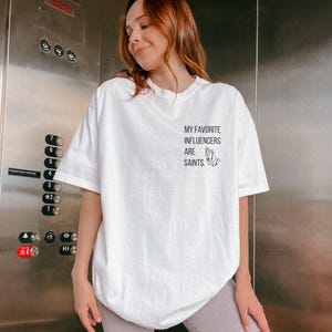 May include: White t-shirt featuring the text "MY FAVORITE INFLUENCERS ARE SAINTS" and a graphic of praying hands. The shirt has a loose fit and short sleeves. The word "White" is printed at the bottom of the image.