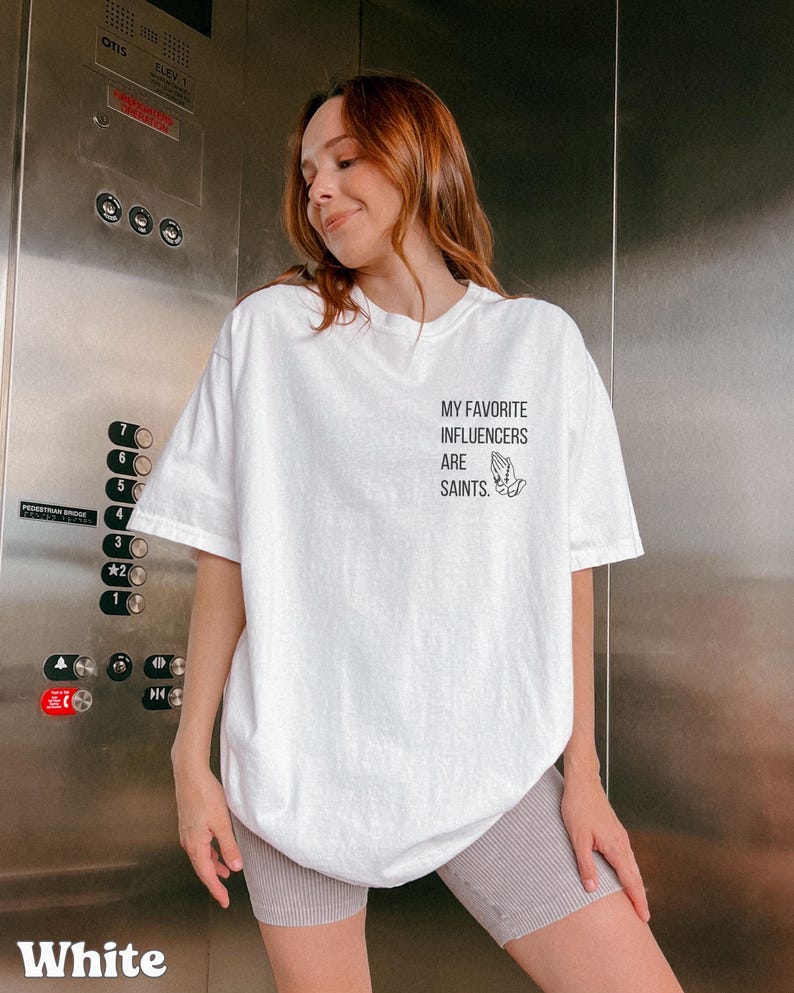 May include: White t-shirt featuring the text "MY FAVORITE INFLUENCERS ARE SAINTS" and a graphic of praying hands. The shirt has a loose fit and short sleeves. The word "White" is printed at the bottom of the image.
