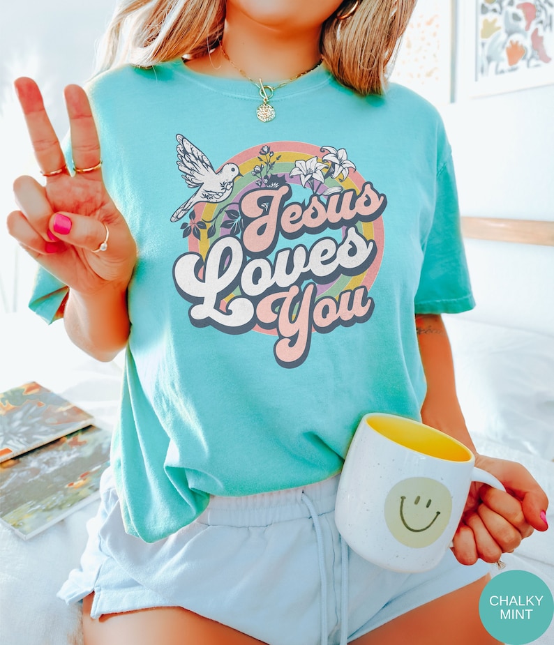 May include: A turquoise blue t-shirt with a retro rainbow design and the text "Jesus Loves You".