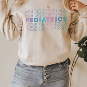 K&ouml;nnte beinhalten: Ein hellbeiger Pullover mit dem Wort "PEDIATRICS", das in einem Regenbogen-Farbverlauf wiederholt wird. Der Pullover wird von einer Person mit langen blonden Haaren und einer blauen Jeans getragen.