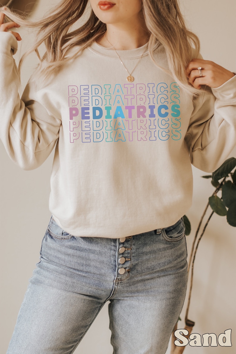 K&ouml;nnte beinhalten: Ein hellbeiger Pullover mit dem Wort "PEDIATRICS", das in einem Regenbogen-Farbverlauf wiederholt wird. Der Pullover wird von einer Person mit langen blonden Haaren und einer blauen Jeans getragen.