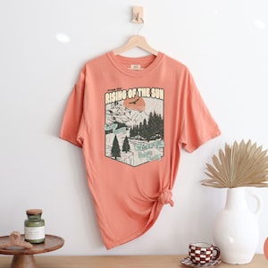 May include: A coral coloured t-shirt with a vintage graphic print of a mountain scene with a sun and the text "From the Rising of the Sun".
