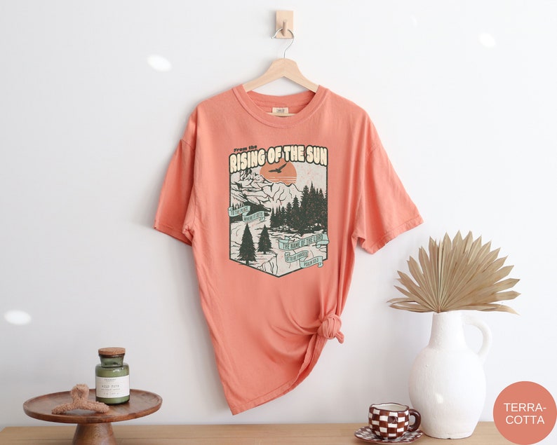 May include: A coral coloured t-shirt with a vintage graphic print of a mountain scene with a sun and the text "From the Rising of the Sun".