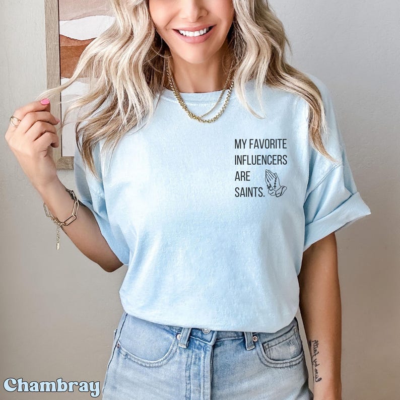 May include: Pale blue t-shirt with rolled sleeves. The shirt displays the text "MY FAVORITE INFLUENCERS ARE SAINTS" in black, accompanied by a graphic of praying hands. The fabric appears soft and comfortable.