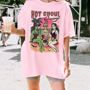 May include: Pale pink t-shirt featuring the words "HOT GHOUL Summer" and a graphic of three cartoon figures on a beach. The figures include a witch, a mummy, and a woman in a swimsuit. The word "Blossom" is printed at the bottom.
