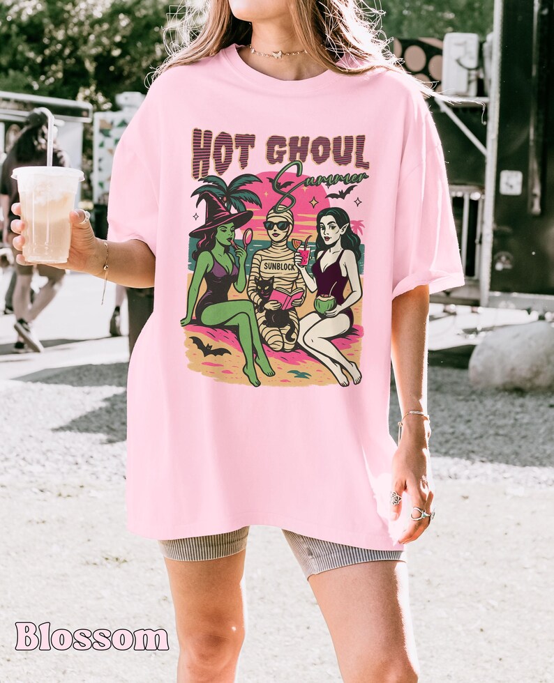 May include: Pale pink t-shirt featuring the words "HOT GHOUL Summer" and a graphic of three cartoon figures on a beach. The figures include a witch, a mummy, and a woman in a swimsuit. The word "Blossom" is printed at the bottom.