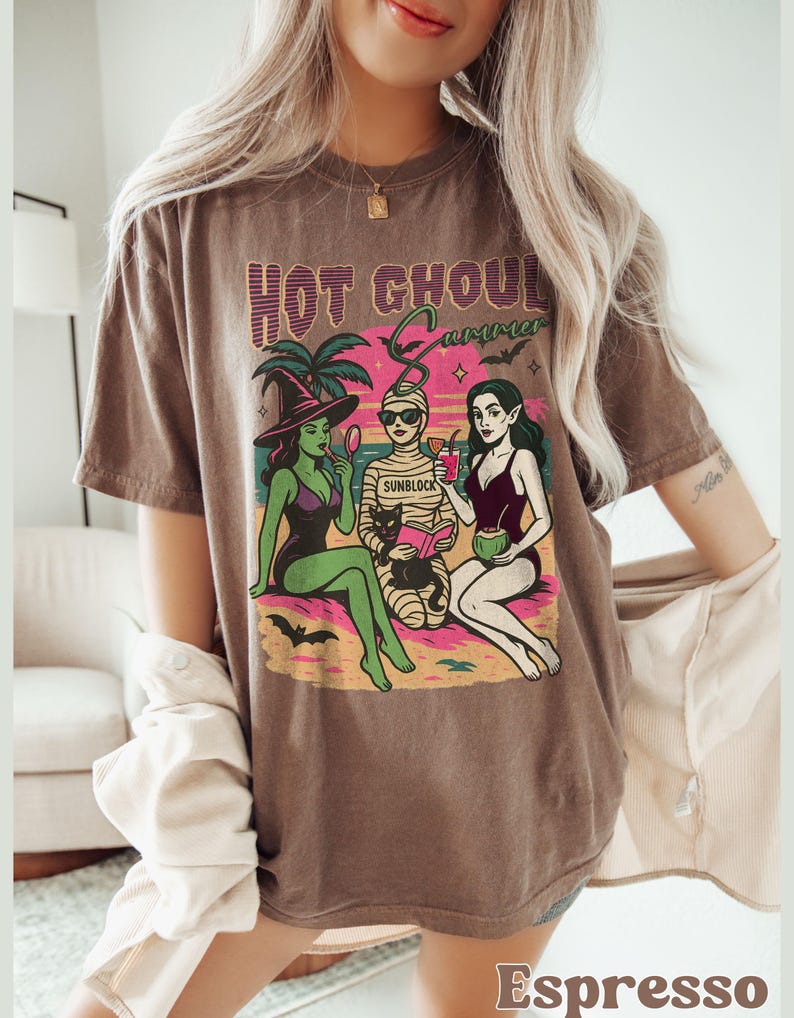 A model wears an espresso color shirt with a retro summerween graphic of a witch, a mummy, and a woman with elf pointy ears sitting on a beach with a black cat in front os a sunset and a palm tree. It says Hot Ghouls summer above them with some bats flying around.