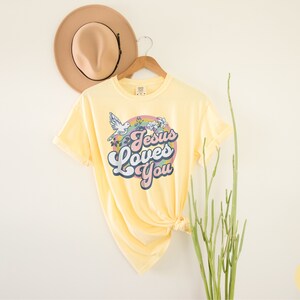 May include: A yellow t-shirt with a retro-style graphic that says "Jesus Loves You" in a rainbow-colored circle with flowers and a dove.