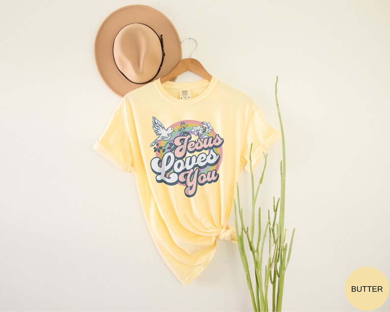 May include: A yellow t-shirt with a retro-style graphic that says "Jesus Loves You" in a rainbow-colored circle with flowers and a dove.