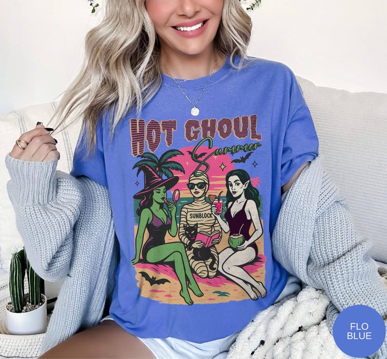 May include: Blue t-shirt with a graphic of three cartoon ghouls on a beach, with the text "HOT GHOUL SUMMER". One ghoul is a witch, another a mummy, and the third a vampire. The shirt is a solid blue colour.
