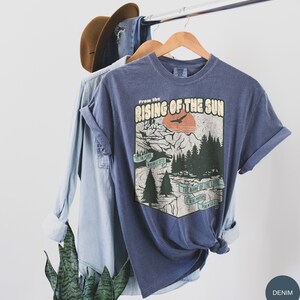 May include: A blue t-shirt featuring a vintage-style graphic of a mountain scene with a sunset and the text "From the Rising of the Sun" and a quote from Psalm 113:3. The shirt is on a hanger with a denim shirt and a brown hat in the background.