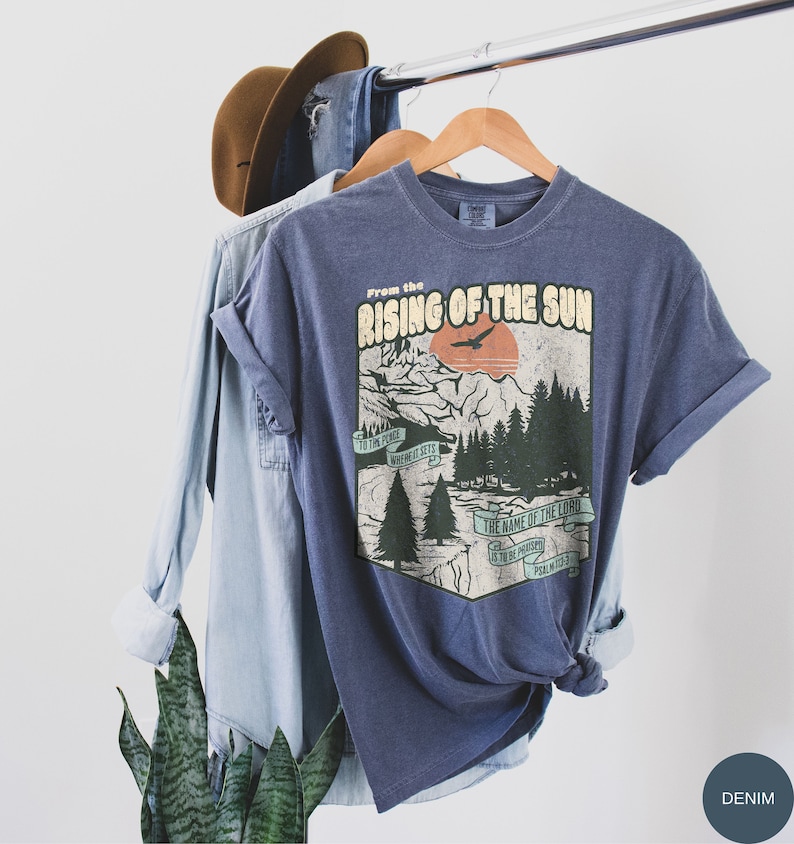 May include: A blue t-shirt featuring a vintage-style graphic of a mountain scene with a sunset and the text "From the Rising of the Sun" and a quote from Psalm 113:3. The shirt is on a hanger with a denim shirt and a brown hat in the background.