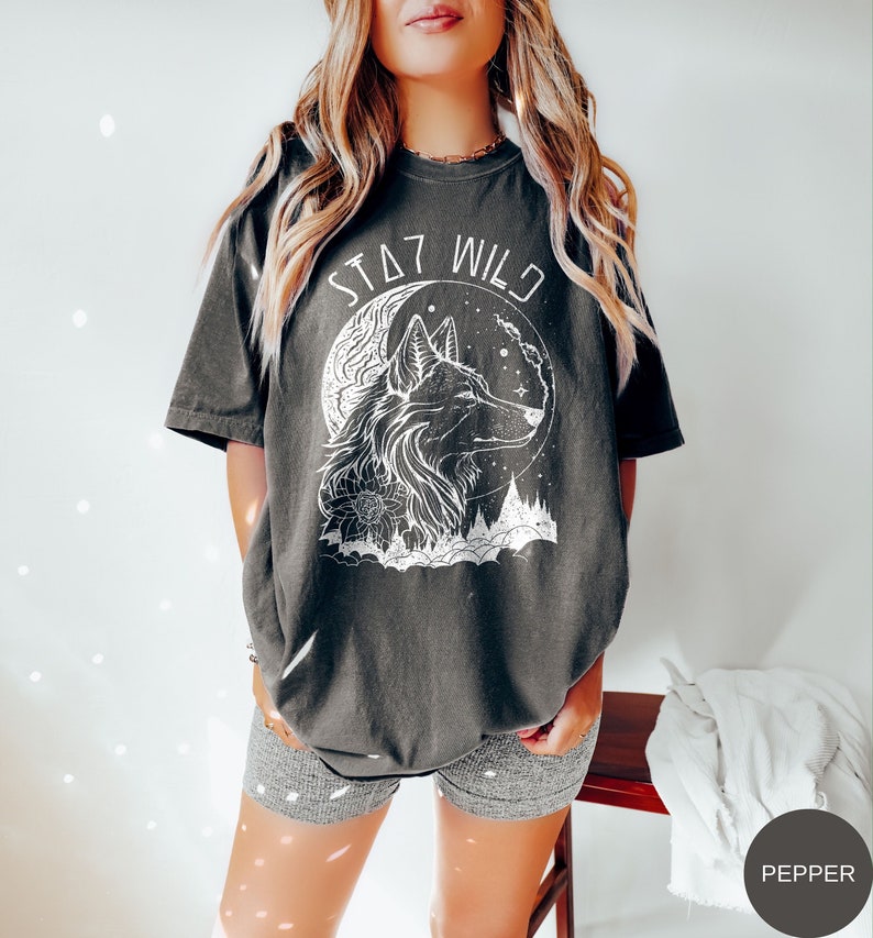 A model wearing a pepper colored shsirt with an outline graphic of a wolf with a crescent moon behind it and some flowers and trees beneath. The words Stay Wild in a futuristic font are written above.
