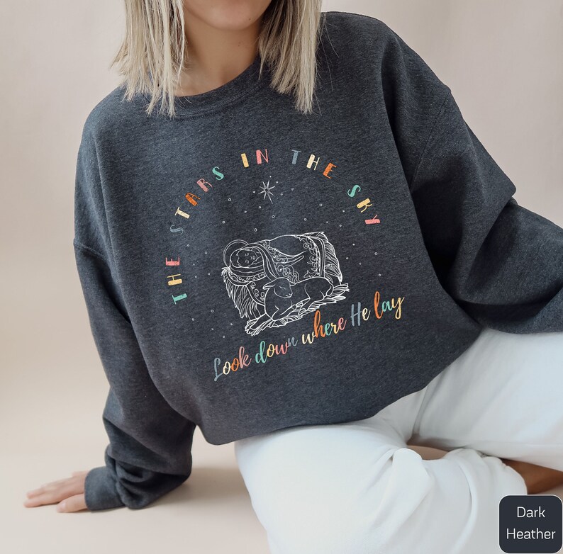 A model is wearing a dark heather sweatshirt and it has a graphic of an outline of baby Jesus and a lamb sleeping under the stars. It says the stars in the sky look down where He lay. The letter colors are alternating muted rainbow colors.