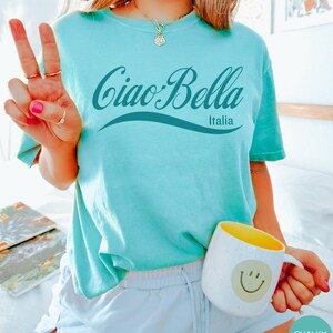 A model wears a chalky mint shirt that says ciao-bella is a classic script style. It has a wavy ribbon underneath and says Italia.