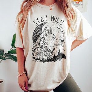 May include: A cream colored t-shirt with a black and white graphic of a wolf's head in front of a crescent moon. The text "STAY WILD" is printed above the wolf.