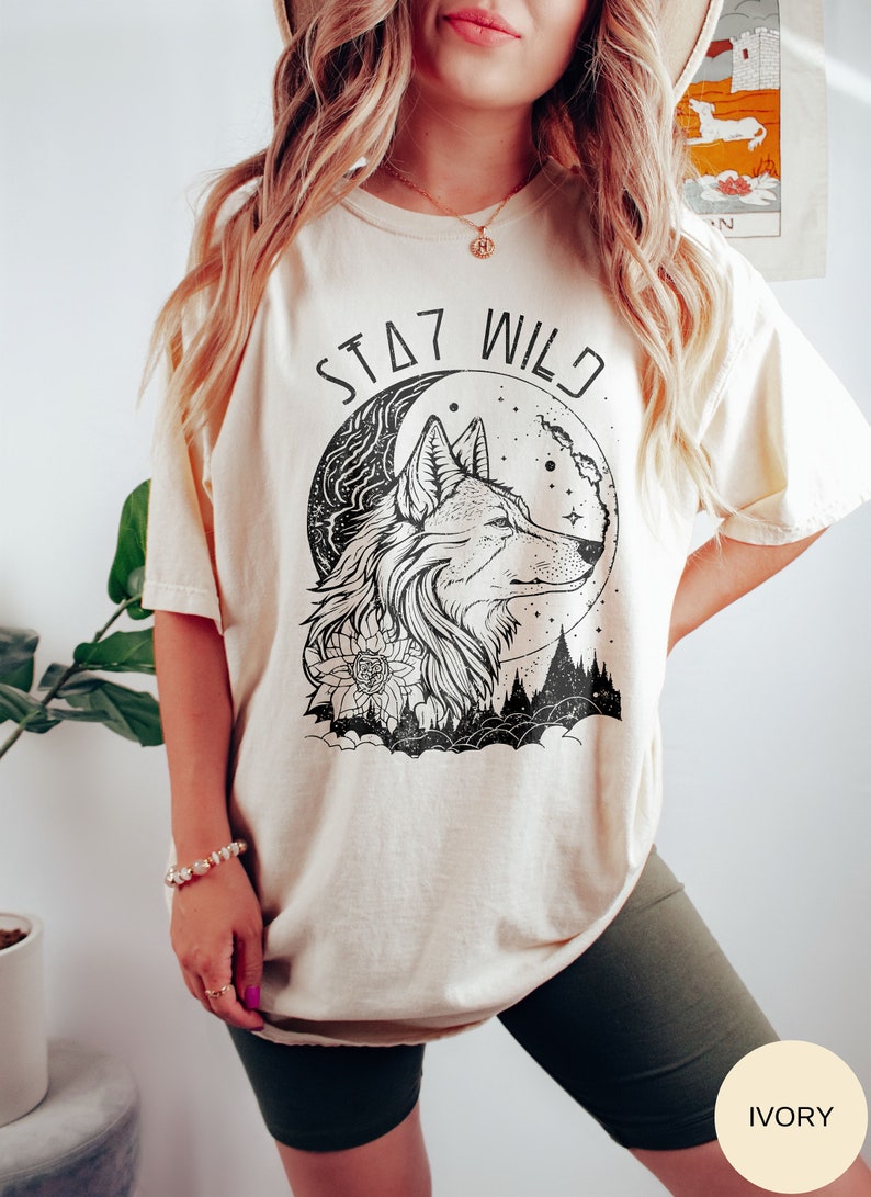 May include: A cream colored t-shirt with a black and white graphic of a wolf's head in front of a crescent moon. The text "STAY WILD" is printed above the wolf.