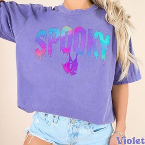 May include: A violet-colored t-shirt with the word "SPOOKY" in a dripping, rainbow-colored font. A bat graphic is below the word. The shirt has short sleeves and a crew neck.