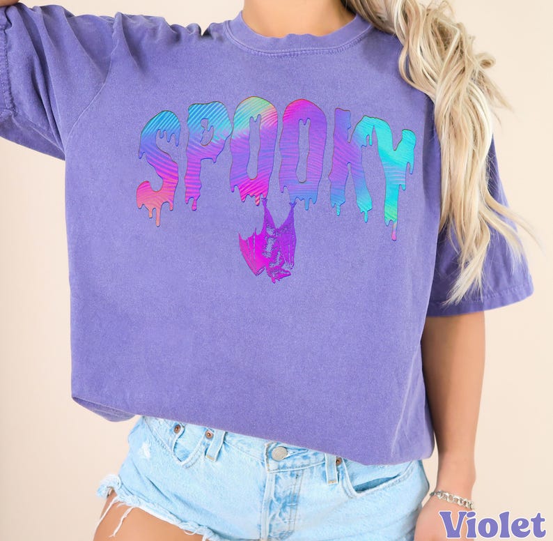 May include: A violet-colored t-shirt with the word "SPOOKY" in a dripping, rainbow-colored font. A bat graphic is below the word. The shirt has short sleeves and a crew neck.