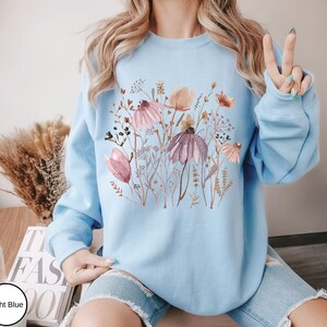 May include: A light blue sweatshirt with a floral print featuring pink, brown, and white flowers. The flowers are arranged in a cluster and are surrounded by green leaves.