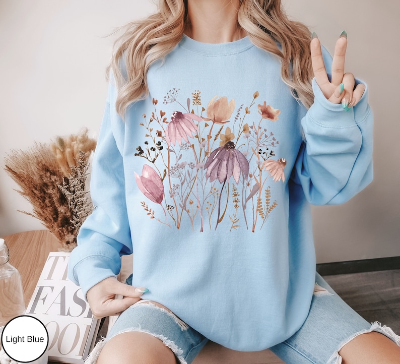 May include: A light blue sweatshirt with a floral print featuring pink, brown, and white flowers. The flowers are arranged in a cluster and are surrounded by green leaves.