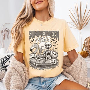 May include: A light yellow t-shirt with the words "Summerween Vibes" in a spooky font. The graphic features a skeleton relaxing on a flamingo floatie, surrounded by jack-o'-lanterns and bats. The shirt is a comfortable fit.