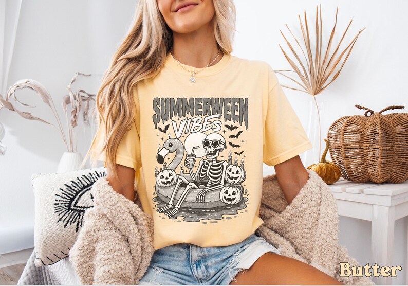 May include: A light yellow t-shirt with the words "Summerween Vibes" in a spooky font. The graphic features a skeleton relaxing on a flamingo floatie, surrounded by jack-o'-lanterns and bats. The shirt is a comfortable fit.