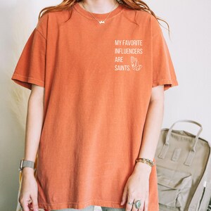 May include: Burnt orange t-shirt featuring the text "MY FAVORITE INFLUENCERS ARE SAINTS" and a praying hands graphic. The shirt has a relaxed fit and short sleeves. The material is soft.