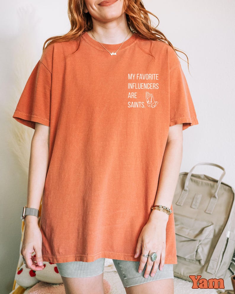 May include: Burnt orange t-shirt featuring the text "MY FAVORITE INFLUENCERS ARE SAINTS" and a praying hands graphic. The shirt has a relaxed fit and short sleeves. The material is soft.