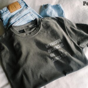 May include: A dark grey t-shirt with the text "MY FAVORITE INFLUENCERS ARE SAINTS" in white, and a graphic of praying hands. The shirt is paired with light blue denim jeans.
