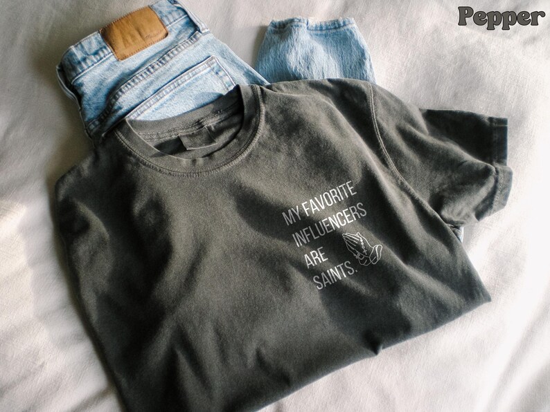 May include: A dark grey t-shirt with the text "MY FAVORITE INFLUENCERS ARE SAINTS" in white, and a graphic of praying hands. The shirt is paired with light blue denim jeans.